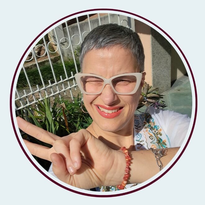 Foto colorida de uma mulher de meia-idade, num dia ensolarado, usando blusa branca estampada de flores e óculos cor marfim. Sorri para a câmera, usando batom vermelho, e tem os cabelos grisalhos bem curtos. Em primeiro plano, faz um sinal de "V" com a mão, cujo punho traz uma pulseira de prata e outra de pedras vermelhas. Ao fundo, Uma grade branca e um canteiro de flores.